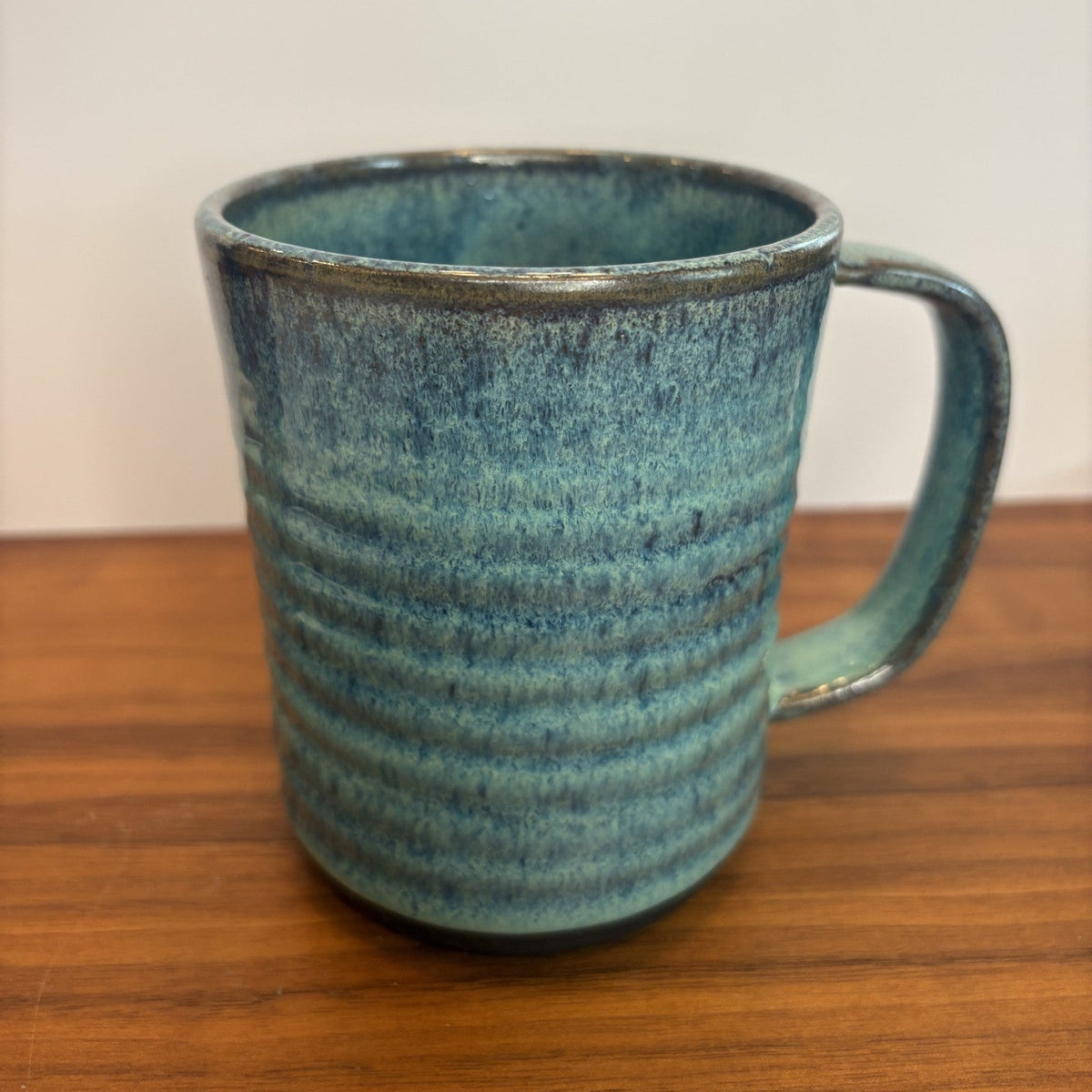 A handmade ceramic mug with a textured, ribbed surface and a C-shaped handle, featuring a mottled glaze of teal, blue, and brown tones. The mug is placed on a wooden tabletop against a plain off-white wall.