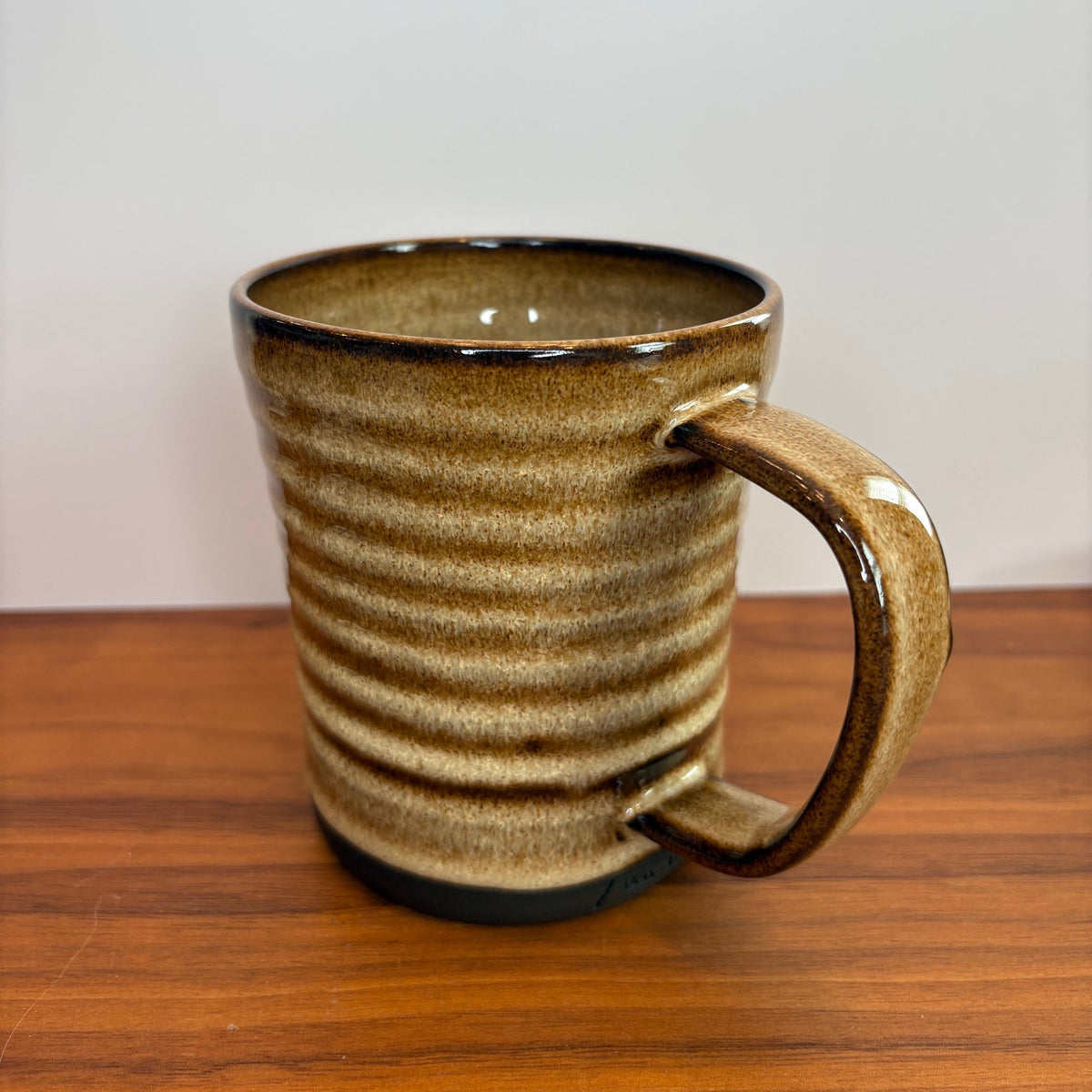 A handmade ceramic mug with a ribbed texture and a curved handle, featuring a speckled brown and beige glaze with a dark brown rim, sitting on a wooden table against a plain light gray wall.